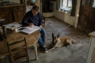 Middle-aged man at a farmhouse kitchen table with a calm Belgian Malinois lying beside him, sunlit rustic interior, subtle connection between them.